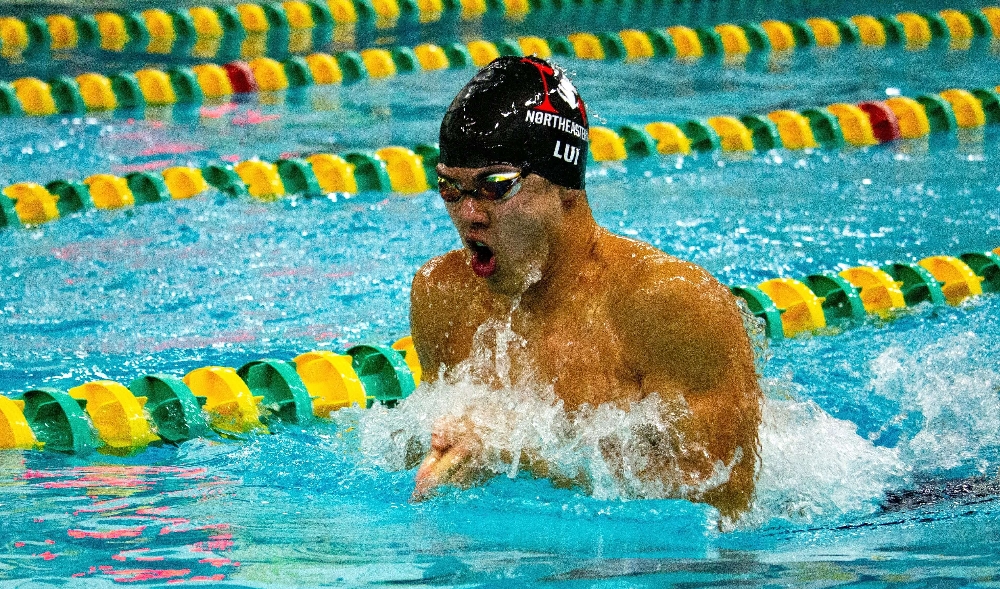 Competitive swimmer performing the breaststroke in a university pool, wearing goggles and a swim cap, with colorful lane dividers in the background.